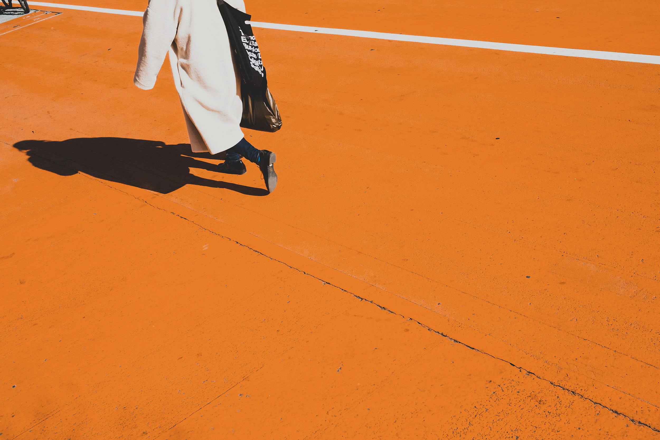 Person in a long beige coat walking across a bright orange pavement, casting a strong shadow, with a black tote bag over their shoulder and a white line marking the ground in the background.