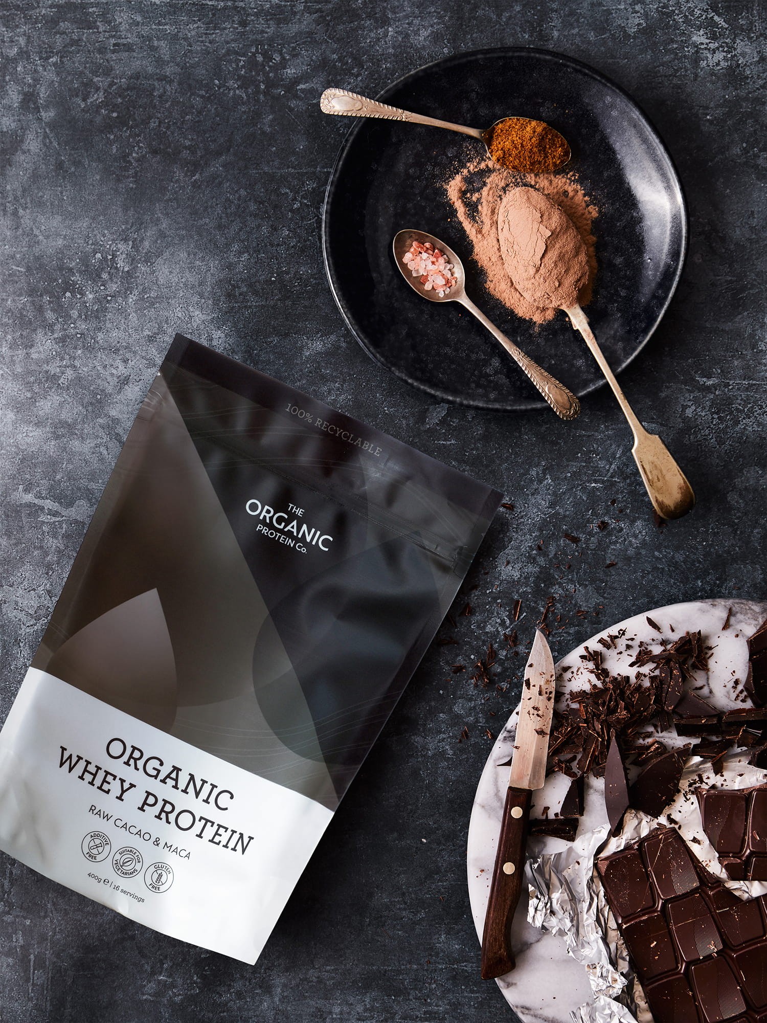 Packaging for The Organic Protein Co organic whey protein (raw cacao & maca) alongside cacao powder, pink salt, and chopped dark chocolate on a dark stone surface.